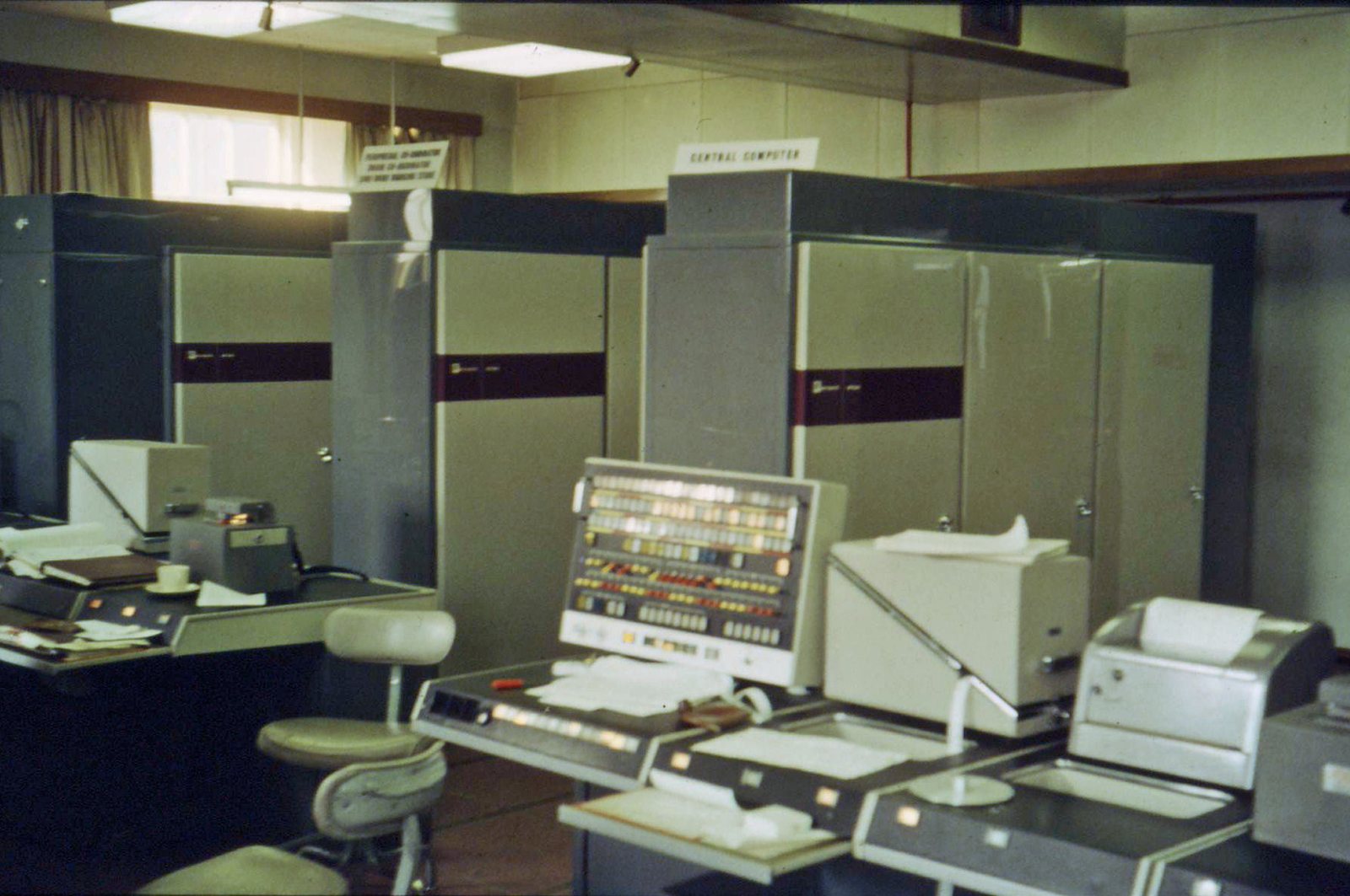 A 1960s computer room: tall grey cabinets labelled central computer, an operator's console covered in switches and lamps, and tape and printer units