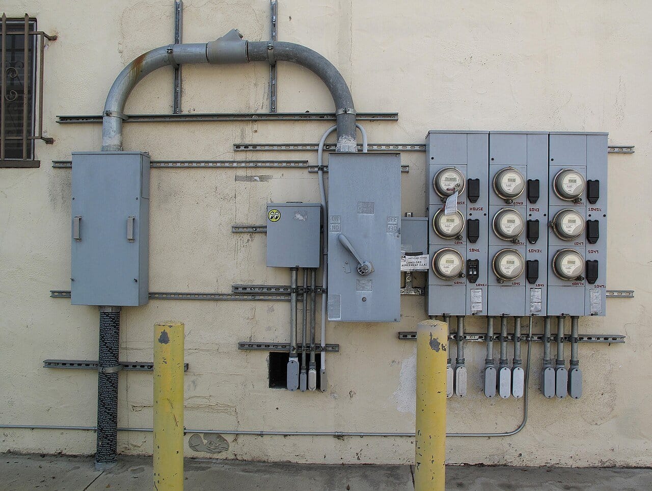 The back wall of an apartment building with one electrical feed running into a bank of nine meters, most labelled with a flat number
