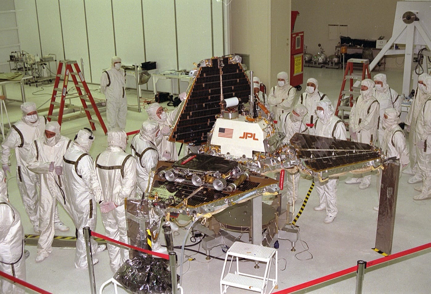 Technicians in white clean-room suits around the Mars Pathfinder lander, a pyramid of solar-panel petals with a small six-wheeled rover on one petal