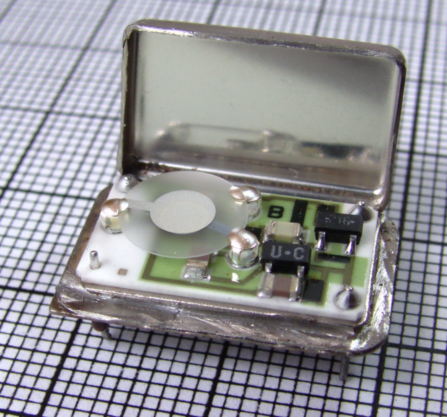 An opened crystal oscillator package on graph paper, showing a thin round quartz disc mounted above a small circuit board
