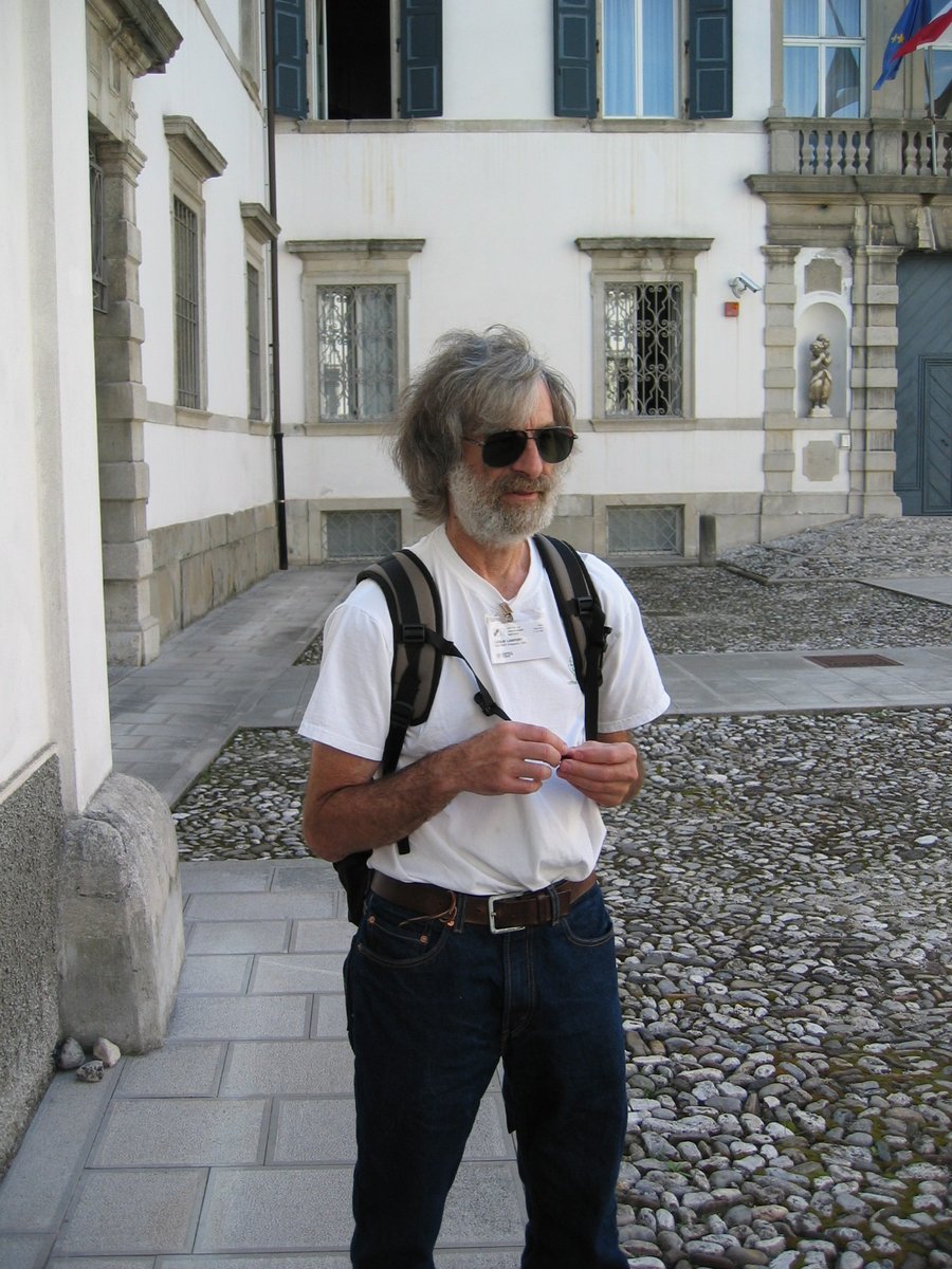 Leslie Lamport, grey-haired and bearded, in sunglasses and a T-shirt, standing in a cobbled courtyard in Udine in 2006