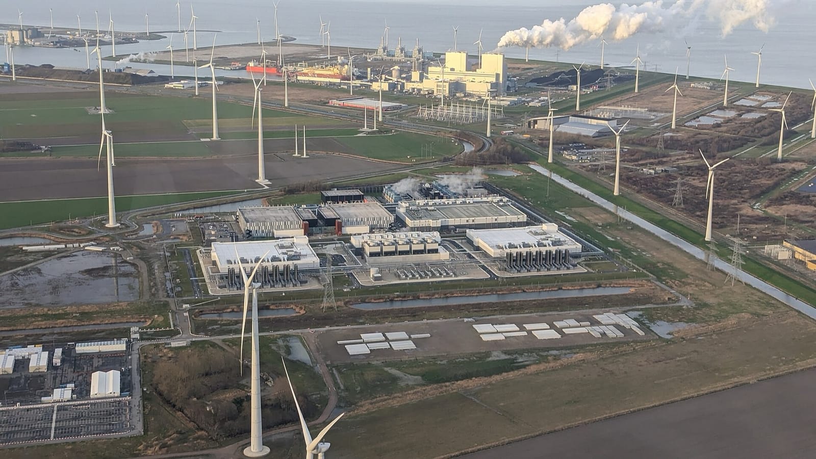 An aerial view of a group of large flat data centre buildings surrounded by wind turbines and farmland