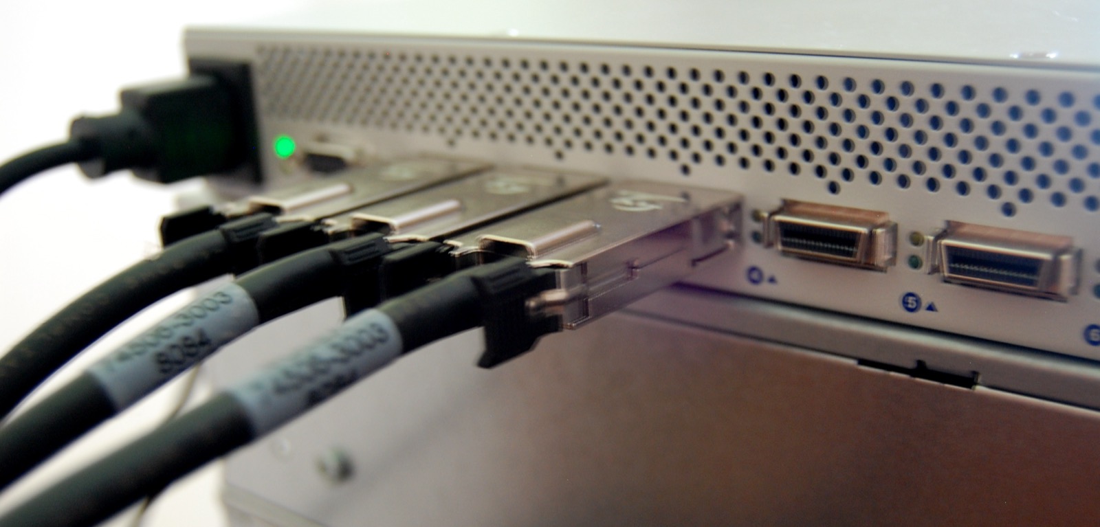 Close-up of the front of a metal network switch with a green status light, three thick black cables plugged into ports, and two empty ports beside them