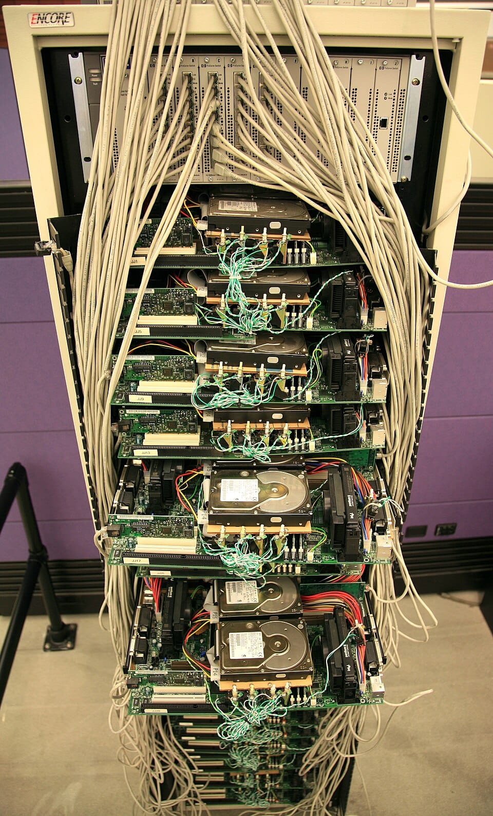 A tall metal rack crammed with bare circuit boards, hard drives and tangled cables, with the boards mounted at angles and partly overlapping