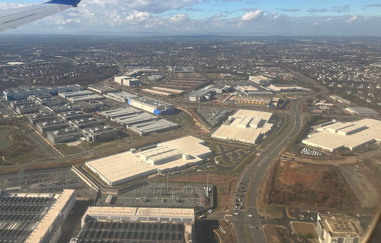 An aerial view of many large, flat-roofed windowless buildings and electrical substations among roads and housing