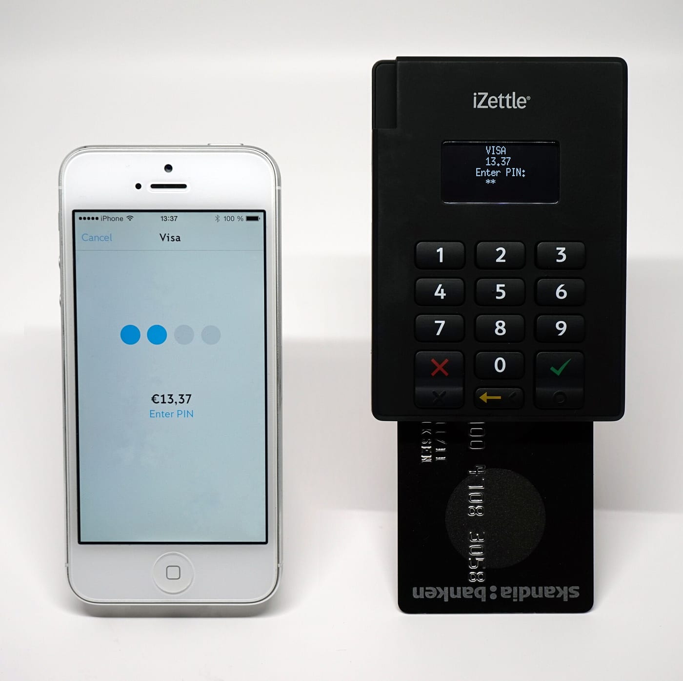 A small card reader with a card inserted, asking for a PIN, next to a phone showing a payment of 13.37 euros in progress