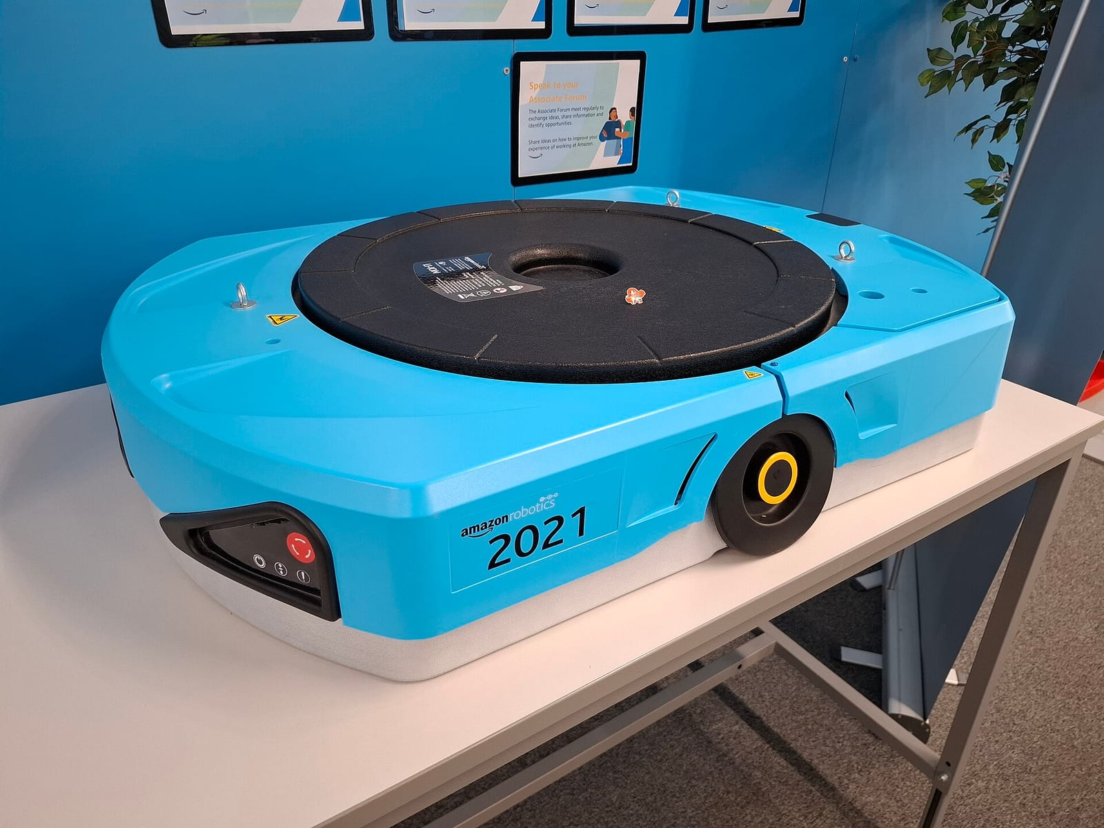 A low, round-edged blue robot with two wheels and a black turntable on top, on display on a table