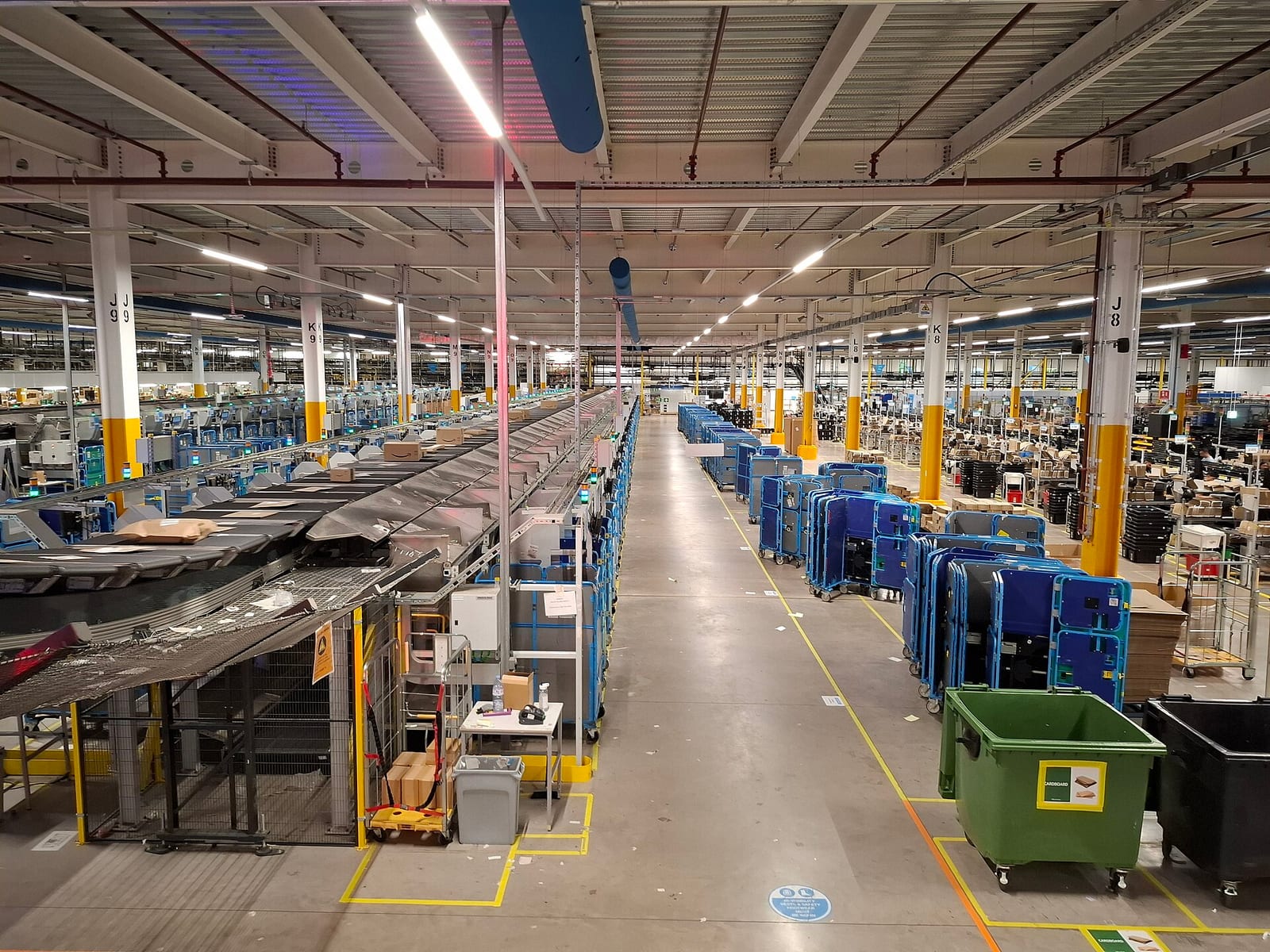 A huge warehouse floor seen from above, with a long conveyor carrying boxes on the left and rows of blue roll cages on the right