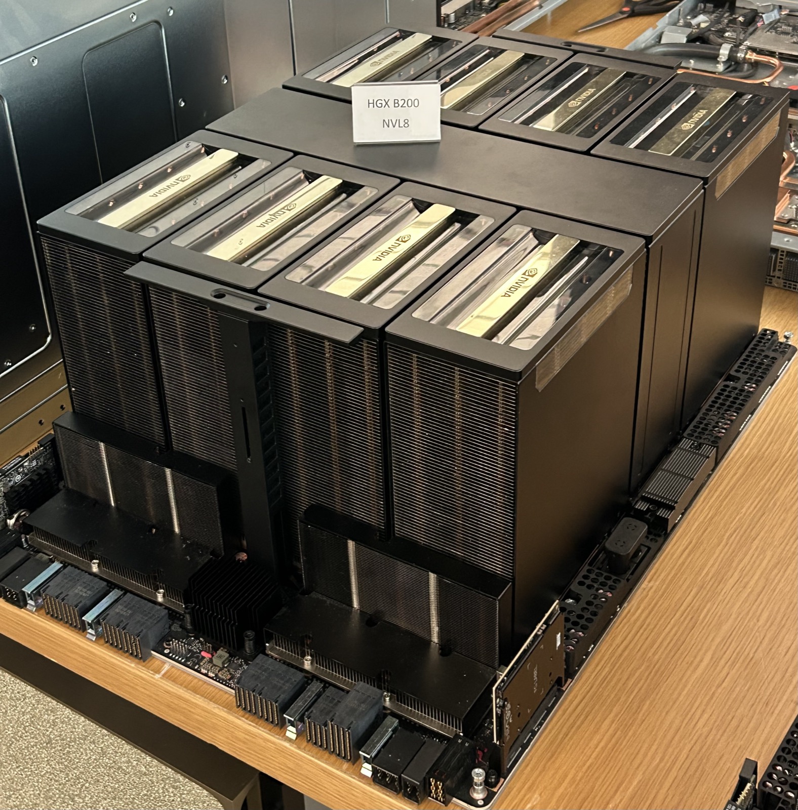 An NVIDIA HGX B200 board on a table: eight GPU modules under tall black heatsinks in two rows of four, with a placard reading HGX B200 NVL8
