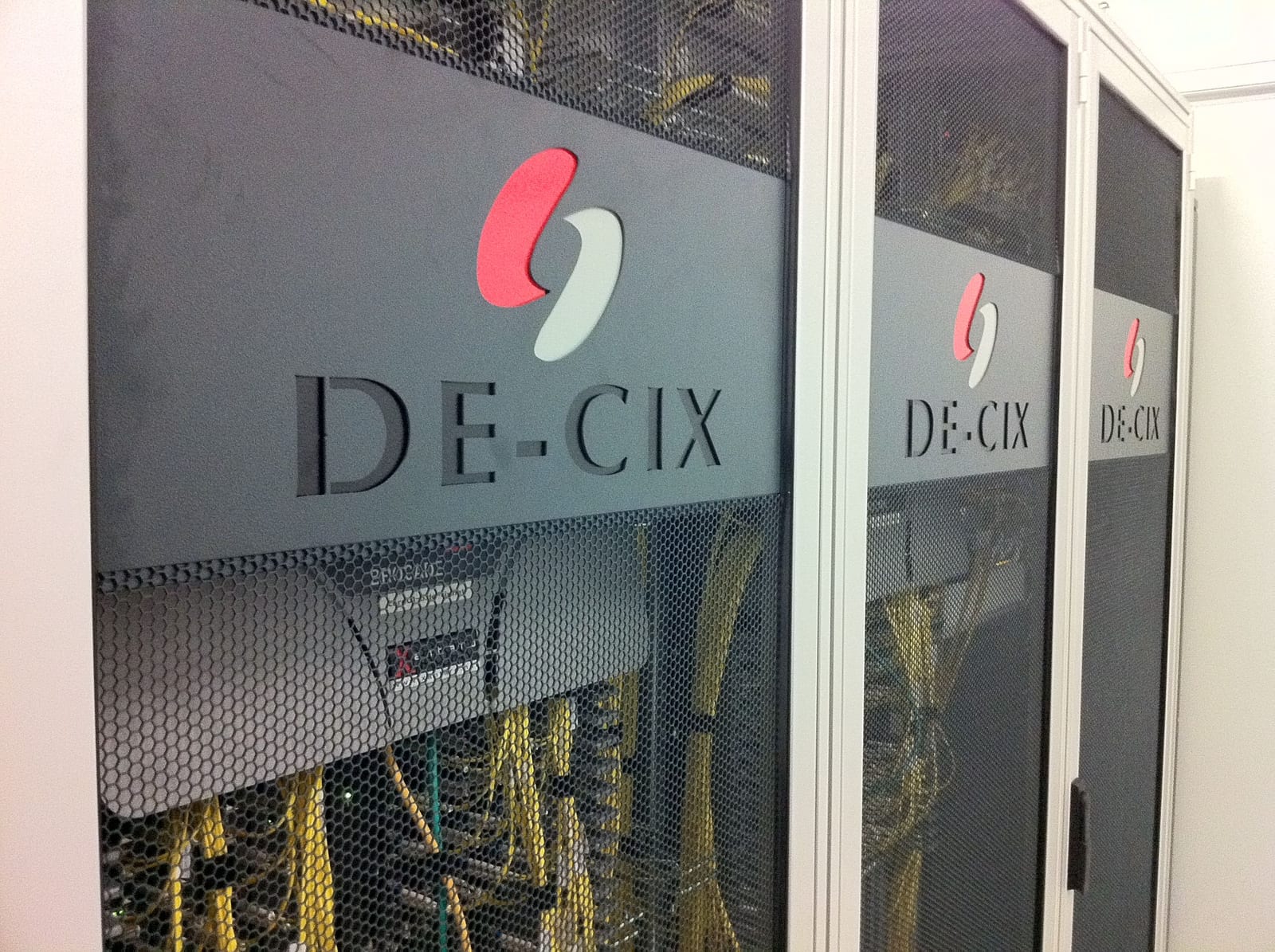 Three black cabinets with perforated doors, each with the DE-CIX logo cut into it, and yellow fibre cords visible behind