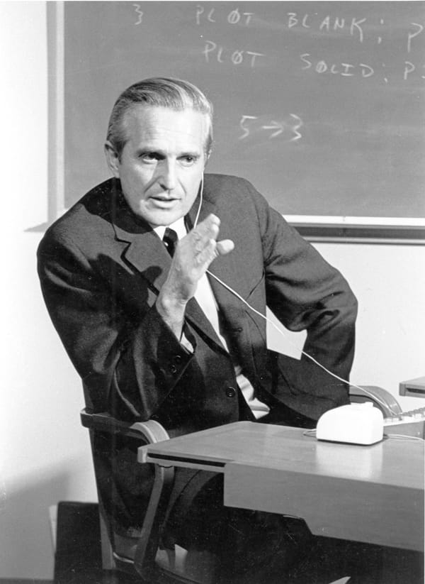 Douglas Engelbart seated at a console with a keyboard, a five-key chord keyset and a mouse, wearing a headset