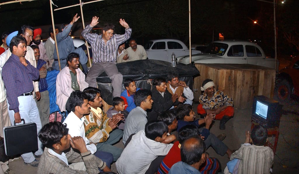 A crowd gathered at night around a small television showing a cricket match, one man cheering with his arms raised