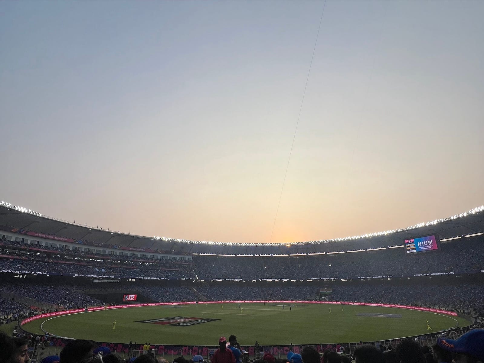A full cricket stadium at dusk with the floodlights on, seen from the stands