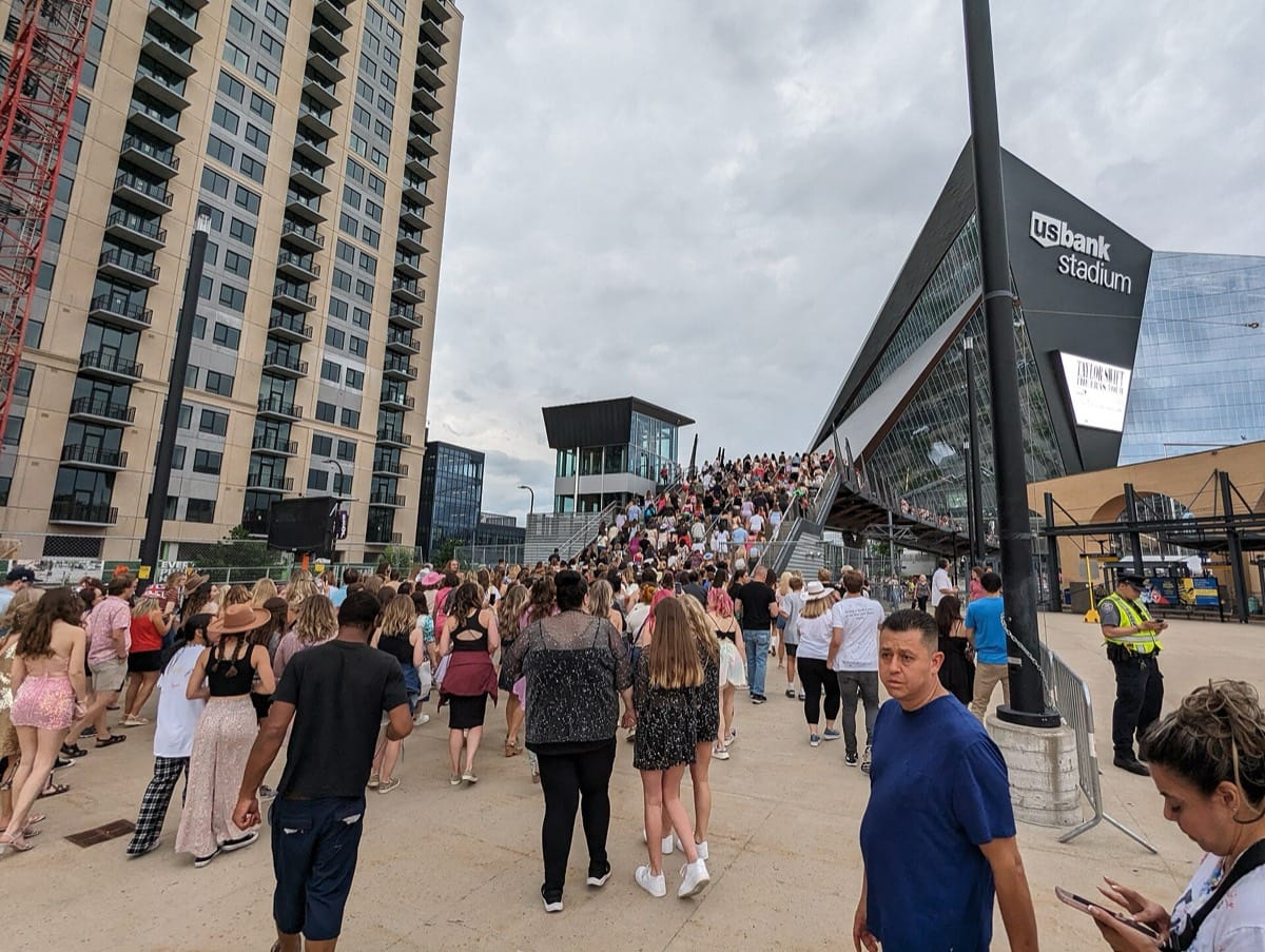 A large crowd of fans, many in sequins and cowboy hats, walking up a ramp towards a glass-fronted stadium with a sign advertising the Eras Tour