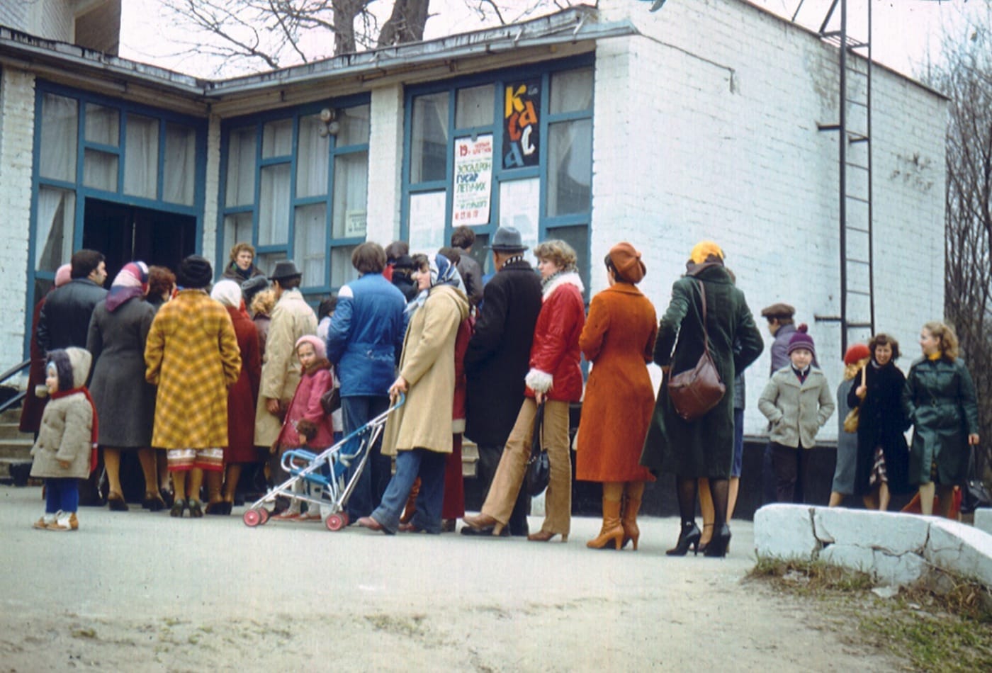 A line of people in winter coats outside a small white building with a sign reading kassa, the Russian for ticket office