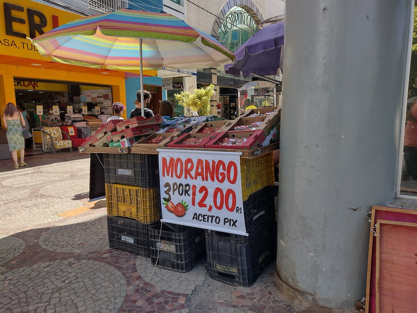 A street fruit stall in Brazil under umbrellas with a hand-written sign reading Morango 3 por 12,00 R$, Aceito Pix