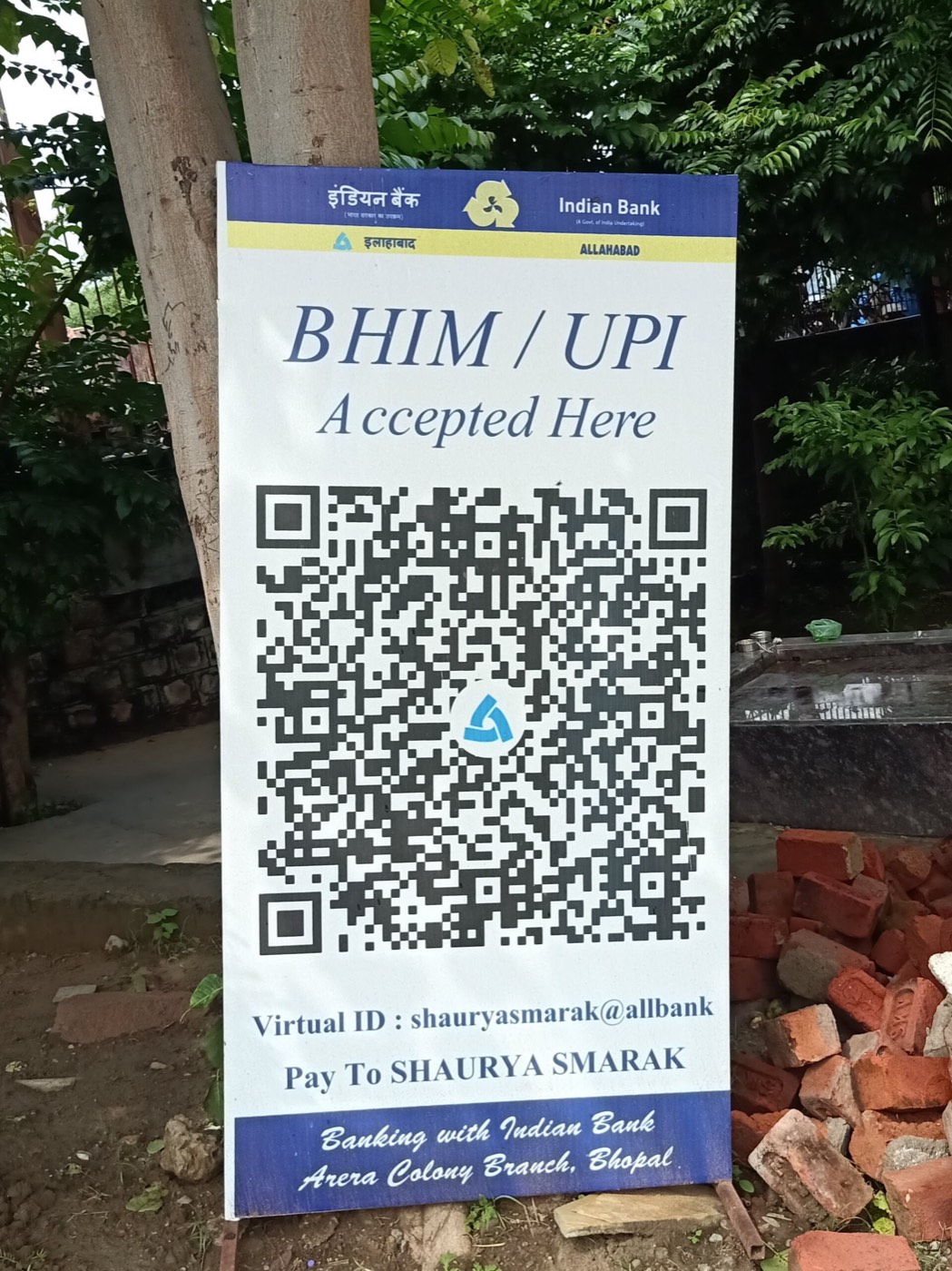 A standing sign reading BHIM / UPI Accepted Here with a large QR code, the Virtual ID shauryasmarak@allbank and Pay To SHAURYA SMARAK, from Indian Bank