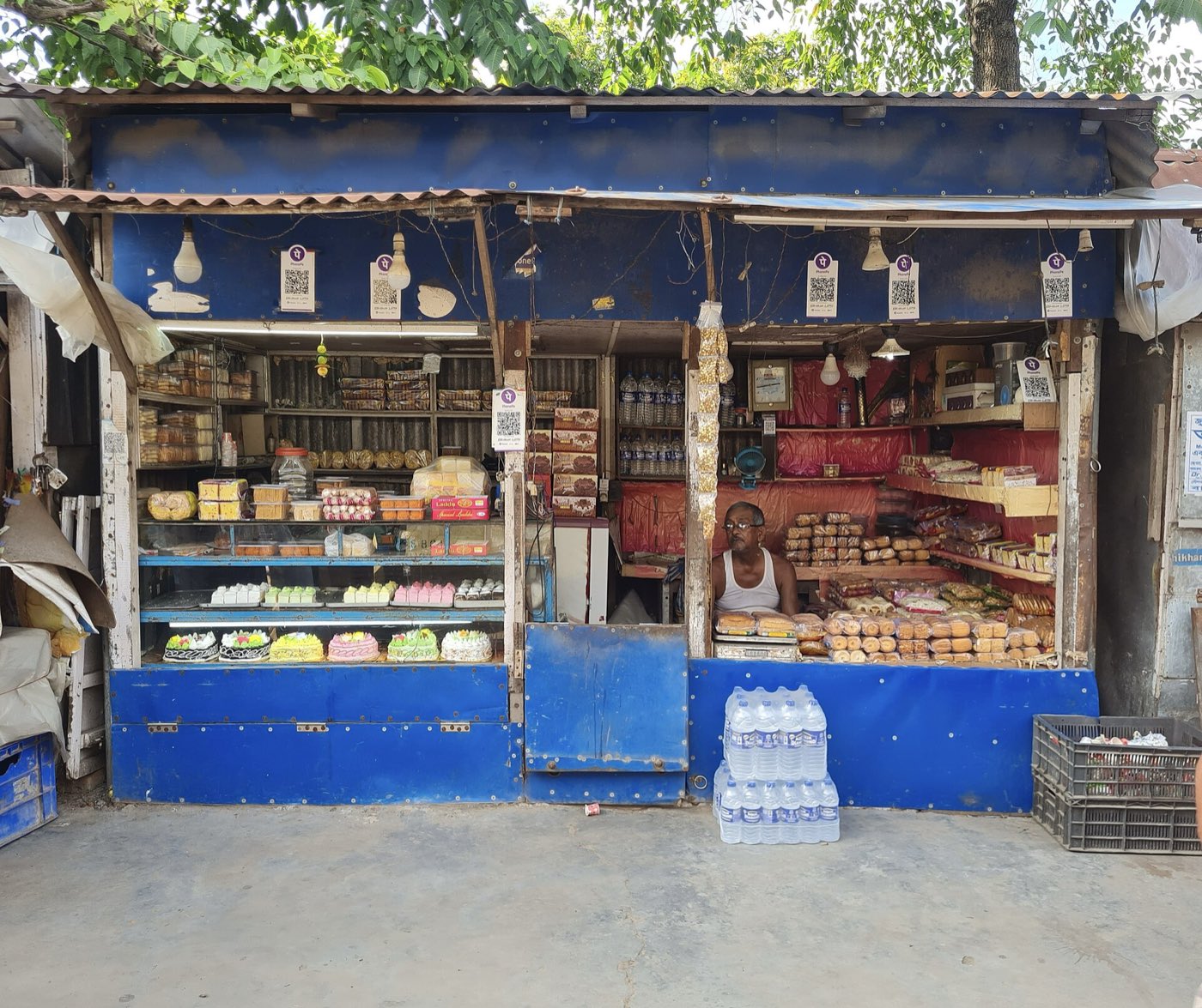 A small roadside sweet shop with a blue tin front, a shopkeeper at the counter, and several purple PhonePe QR code cards hanging around the shop