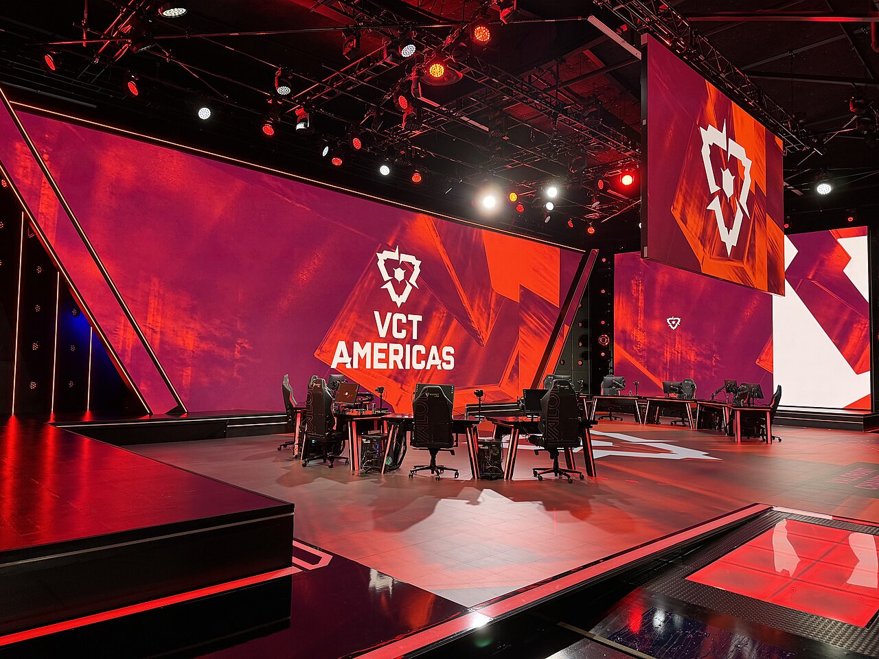 An empty esports stage with two rows of desks, gaming chairs and PCs in front of a large red LED wall