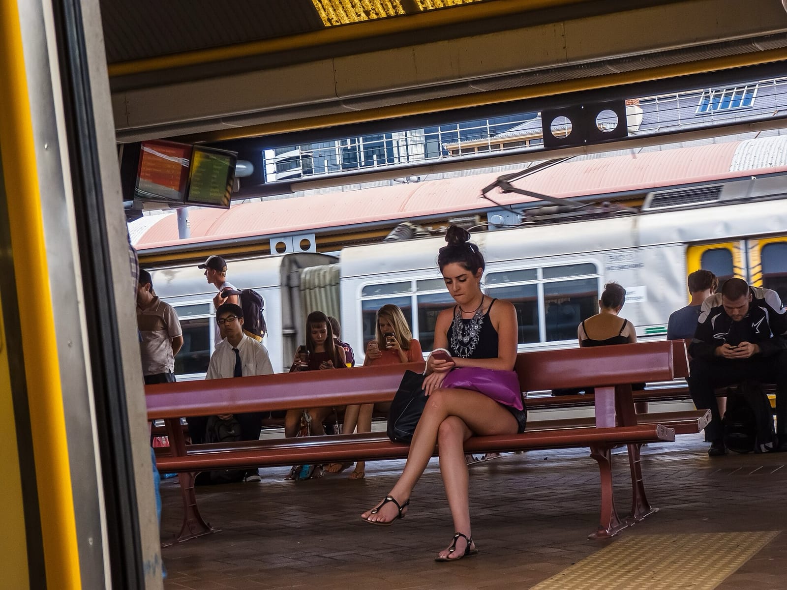 Commuters sitting on benches on a railway platform, most of them looking at their phones, with a train behind them
