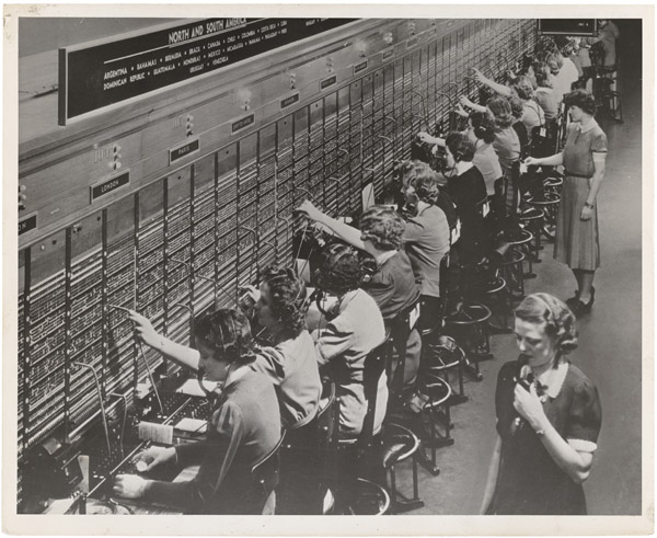 A row of telephone operators with headsets plugging cords into a long switchboard labelled with destinations such as London and Paris
