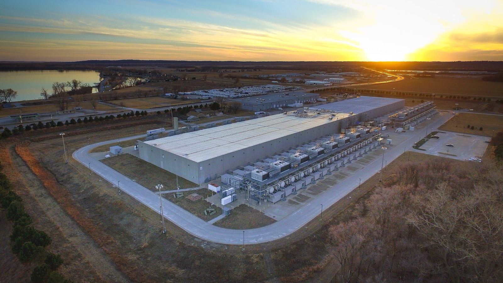 Aerial view of a large, low warehouse-like building with rows of equipment along its side, at sunset