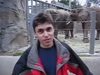 A low-resolution video still of a young man standing in front of an elephant enclosure