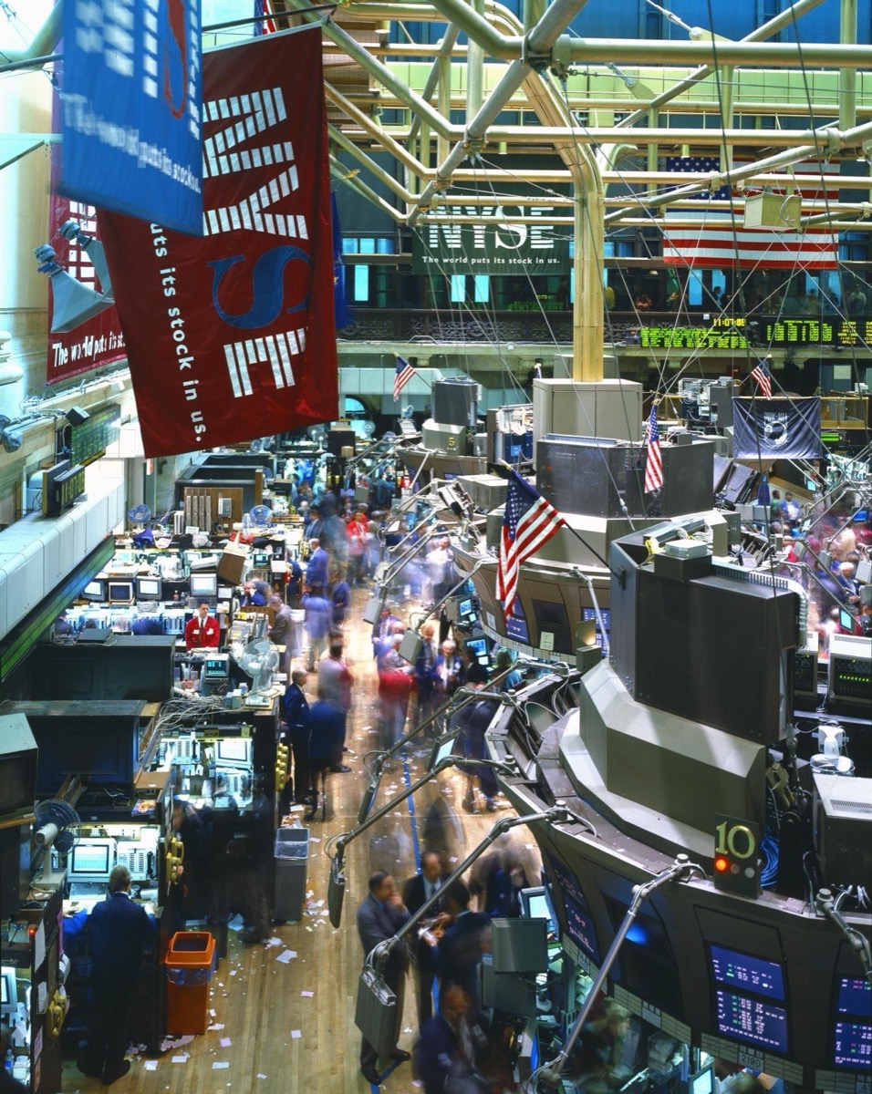 A crowded stock exchange trading floor with traders gathered around trading posts under banks of screens
